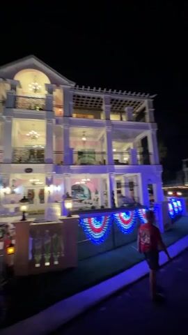 The video begins with a wide shot of a large, multi-story house decorated with white columns and ornate balconies. The house is illuminated with various lights, including chandeliers and string lights. In front of the house, a fence is adorned with red, white, and blue bunting lights, and on the lawn, there are decorative lamps. To the left, a white horse-drawn carriage, also lit with string lights, is parked on the grass. A person in a red shirt and dark shorts is visible on the right side of the frame, walking away from the camera. As the video progresses, the camera pans across the scene, offering different angles of the house and the illuminated carriage. Several palm trees are visible in the background, and the overall setting appears to be a residential area at night.