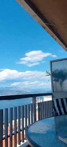 The video shows a view from a balcony overlooking a body of water and distant mountains under a clear blue sky with scattered white clouds. A railing runs along the balcony, and a table with a glass top is partially visible in the foreground. To the right, a chair with black and white striped cushions is partially visible, along with a screen or panel. A palm frond casts a shadow on the screen. The overall impression is of a scenic, outdoor setting, possibly a resort or a home with a view.