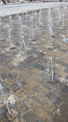 The scene features a water fountain in a public square. The fountain consists of numerous small jets of water spouting up from square stone tiles on the ground. Several people are visible in the background, including people sitting on a bench and a person on a bicycle. A large, old building is also visible in the background. The day appears to be sunny.