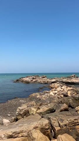 This vertical, eye-level shot captures a serene coastal scene in Orpesa, Spain, under a clear blue sky, suggesting a pleasant day. The foreground is dominated by a rugged, rocky shoreline, with large, weathered stones of various earthy tones, predominantly brown and tan, jutting out into the sea. Some of these rocks appear smooth, worn by the waves, while others have a more textured surface.

Beyond the immediate rocks, the turquoise sea stretches towards the horizon, meeting the sky in a soft, hazy line. Scattered rocky outcrops emerge from the water, creating a natural, irregular breakwater. Small waves gently lap against the rocks, producing white foam and adding a sense of movement to the otherwise still scene. A small amount of dark seaweed or debris is visible in the calmer waters near the shore.

In the distance, on one of the further rocky islands, a tiny figure can be seen, possibly a person standing or observing the scenery. There is no discernible text visible in the image. The overall mood is peaceful and tranquil, highlighting the natural beauty of the Spanish coast on a bright, sunny day. The composition, with the rocks in the foreground leading the eye towards the expansive sea and sky, creates a sense of depth and invites the viewer to immerse themselves in the calm coastal environment.