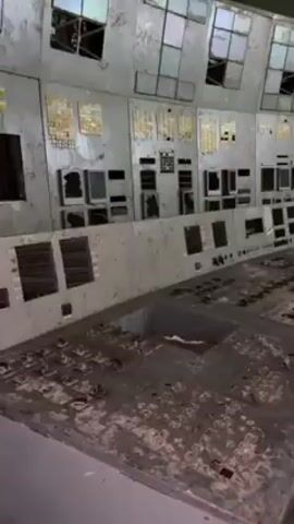 The video shows a large, derelict control room with a curved control panel. The panel is gray and damaged, with missing buttons and a surface covered in debris. The wall behind the panel is lined with square windows, many of which are broken. The room appears abandoned and in disrepair. A beeping sound is present throughout the video.