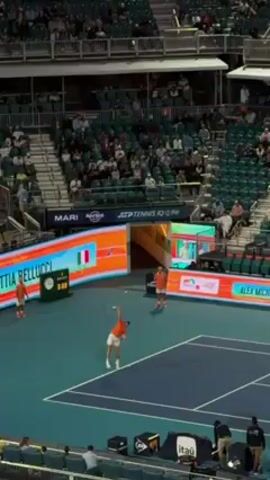 The video shows a tennis match taking place at an outdoor stadium, with a large audience seated in the stands. Two male tennis players, wearing orange outfits, are positioned on a blue tennis court, one is about to serve. Electronic scoreboards display player names "MATTIA BELLUCCI" and "ALEX MICHELSEN" along with a flag. Advertisements for various companies like "ITAÚ" and "Hard Rock" are visible on the court and screens. Several individuals, likely staff or officials, are present around the court. The environment suggests a professional tennis tournament setting.