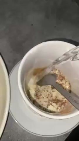 The video shows someone pouring a stream of hot water into a white coffee mug that contains a powder-like substance, a small amount of a brown liquid, and a stainless steel spoon. The water mixes with the contents, creating a light brown liquid with bubbles on the surface. The mug is on a white saucer, and a blurred white dish is partially visible to the left of the mug. The surface underneath appears to be grey.