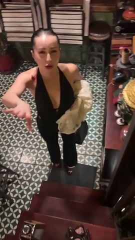 A woman with dark hair pulled back and wearing a black halter top and black pants stands on a step in a room with a patterned tile floor. She is holding a light-colored shawl over her right arm and a dark bag in her left hand. She is looking at the camera and gesturing with her right hand, as if speaking. Her left hand briefly touches her forehead. She smiles at the end of the clip.