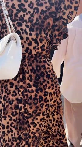 A rear view of a woman wearing a leopard-print dress and a white quilted purse with a gold chain strap walks through a crowded outdoor market. She is followed by a person with dark hair. In the background, other market-goers are visible, along with stalls and a building with signage that appears to be in Cyrillic. The lighting suggests daytime, and the environment is bright and sunny. The individuals are walking in the same direction, with some interaction implied by their proximity and movement. The scene appears to be a bustling commercial area, likely a street market or shopping complex.