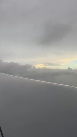 The video shows a view from an airplane window. The foreground is dominated by the airplane's wing, which is a dull gray color. Beyond the wing, the sky is filled with clouds. The clouds are a mix of dark gray and lighter, wispy formations. There are patches of yellowish-white light peeking through the clouds, suggesting the presence of the sun or a break in the cloud cover. The overall lighting is overcast and muted. The scene appears to be captured during the day, likely at some altitude given the presence of clouds. There are no people or distinct objects visible within the frame other than the airplane's wing and the clouds. The camera is stationary, looking out the window.