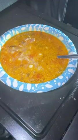 A close-up shot shows a blue and white patterned paper bowl filled with soup. A spoon is submerged in the soup, stirring it. The soup appears to be a hearty broth with small pasta shells, shredded meat, and visible pieces of tomato or other vegetables. Steam rises from the soup, indicating it is hot. The bowl is placed on a dark, flat surface, possibly a stovetop or counter.