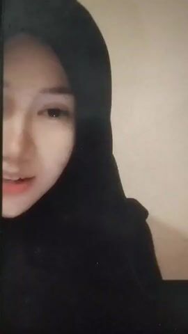 A close-up shot shows the face of a young woman wearing a black hijab. Her eyes are closed, and her head is tilted down. The background is a plain, light-colored wall. The lighting is soft, with some shadows. The woman's expression appears calm or thoughtful.