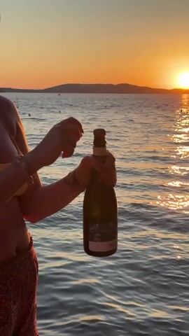 A person's hands are shown in the foreground, uncorking a bottle of champagne. The background features a body of water with ripples reflecting the setting sun, which casts a warm orange glow over the scene. Hills or mountains are visible in the distance on the horizon. The person is wearing a bracelet on their left wrist.