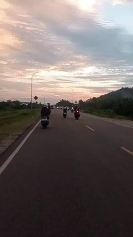 The camera is positioned on the side of a black asphalt road marked with a solid white line. The view looks down the road, following a group of people riding motor scooters in the distance. All riders are wearing helmets. There are several streetlights flanking the road. A yellow road sign with a black symbol is to the left of the road. The horizon is marked by a landscape of scattered buildings and greenery. A hill covered in trees is to the right. The sky above is filled with clouds painted in hues of pale orange and white, indicating either sunrise or sunset.