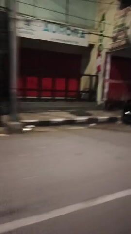 The video is a blurry, shaky first-person perspective of a street scene at night. The camera is moving, possibly on a vehicle. Several motorcycles are parked on the side of the road, and a few people can be seen near them. In the background, there are buildings with closed storefronts and illuminated signs. One scene shows two people riding on a motorcycle. Another shows a food stall with smoke rising from it. The overall impression is that of a busy street in a city or town at night.