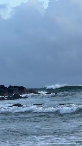 The video captures a dynamic seascape at Thalaramba, Sri Lanka, dominated by the powerful motion of ocean waves under a dramatic, cloudy sky. Large waves crash against dark, rugged rock formations that extend from the left side of the frame into the water. Further out, more significant waves are seen building and breaking, their white crests contrasting with the deep blue-green of the sea. The foreground shows smaller, choppy waves rolling towards the viewer, indicating a rough sea state. The overall weather appears overcast, contributing to the slightly somber yet majestic atmosphere of the scene. No people or text are visible in the video.