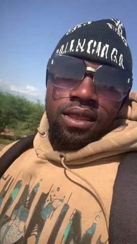The video captures a close-up of a Black man from the chest up, wearing a black beanie with white text and large, dark-tinted sunglasses. He has a beard and is wearing a light brown hooded sweatshirt with a graphic print. A black backpack is visible, with straps going over his shoulders.

The man is looking directly at the camera in some shots and to the side in others, with his head tilted slightly upwards. His expression appears neutral. The scene is outdoors, with a clear blue sky and some scattered clouds visible. The background suggests a dry, somewhat arid environment with sparse vegetation, including some bushes and trees.

The lighting indicates it is daytime, likely late morning or early afternoon, with bright sunlight casting shadows on the man's face. There are no overt signs of any specific activity or event other than the recording of a selfie-style video. The location cues of dry vegetation and a clear blue sky are consistent with an outdoor setting in a warm climate.

The text on the beanie appears to be a brand name, possibly "BALENCIAGA," though it is partially obscured and somewhat stylized. The graphic print on the sweatshirt is abstract and colorful, with elements of blues, greens, and black.