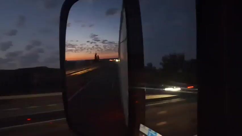 The video shows a view from a vehicle's side mirror at dusk. The mirror reflects a highway with several cars driving on it. The sky is a deep blue with orange and pink hues near the horizon, indicating the sunset or sunrise. There are some clouds in the sky. The landscape on either side of the highway appears to be hilly and sparsely vegetated, with trees visible in the distance. The camera is pointed towards the right side of the vehicle, with the edge of the vehicle's body visible in the foreground on the right. The overall atmosphere is peaceful and serene, capturing the beauty of the twilight hour.
