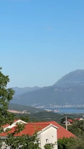 The video pans across a scenic landscape with a clear blue sky. In the foreground, there are rooftops of houses, some with red tiles, interspersed with green foliage and trees. In the mid-ground, there is a body of water, possibly a bay or a lake, with a town or city visible along its shores. Further in the background, majestic mountains rise, their peaks disappearing into the haze. Power lines and poles are also visible, suggesting the presence of infrastructure. The overall impression is one of a peaceful, rural or semi-rural setting on a sunny day.