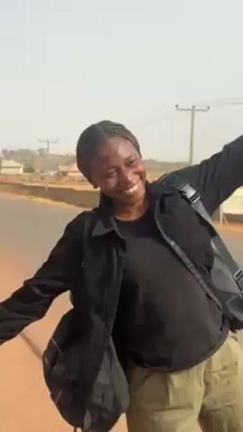 A young woman stands smiling on the side of a road. She wears a black shirt and jacket, olive-colored pants, and has her hair in braids. She carries a large black bag on her left shoulder, and a smaller black bag across her body. She has her arms spread wide at first, then brings them down. She is smiling and appears to be posing for a photo or video. The road behind her has a red dirt shoulder and a paved surface. Houses can be seen in the background. The sky is bright and slightly hazy.