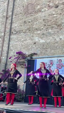 This video captures a group of young people performing a traditional dance on a stage. The dancers, all dressed in matching black dresses with red accents and red headscarves, are positioned against a backdrop of a textured stone wall and a banner. The banner displays images of performers in colorful, elaborate costumes. Purple flowers cascade down from above the banner, adding a splash of color to the scene.

The dancers' costumes consist of black, floor-length dresses with gold trim and detailing around the neckline and sleeves. They wear bright red stockings and dark shoes. Some dancers are also seen holding dark-colored accessories, possibly part of their performance props. Their expressions appear joyful and engaged in the dance.

The stage is set outdoors, with a clear sky suggesting it is daytime, likely late afternoon or early evening given the lighting. The stone wall suggests a historic or outdoor venue. The presence of the banner with images of performers hints at a cultural festival or performance event. The overall atmosphere seems festive and celebratory. No discernible text is visible on the clothing or in the immediate surroundings, beyond the images on the banner. The setting in Calella, Spain, provides geographical context to this cultural display.