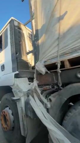 The video shows the side of a large truck from a low angle. The truck is covered by a large, white tarp that casts a striped shadow on the tarp itself. The tarp is secured with ropes. Underneath the tarp, the tops of wooden pallets and some boxes are visible. The truck has dual rear wheels. The scene appears to be outdoors, possibly on a road, with a clear blue sky visible at the top of the frame. The lighting suggests it is daytime, likely in the morning or late afternoon due to the angle of the sun.