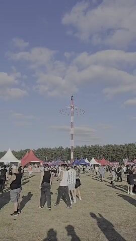The video shows a tall amusement park ride, likely a swing ride, with people riding in chairs that are swinging outwards. The ride is red and white striped and stands against a blue sky with some clouds. In the foreground, there is a grassy field with several tents and a crowd of people. The people are standing around, and some are walking. The overall scene appears to be a festival or fair.