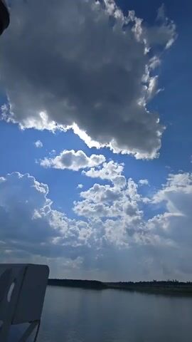 The video captures a view looking up at a bright blue sky filled with various clouds. Some clouds are dark and dense, suggesting potential rain, while others are lighter and wispy, illuminated by sunlight filtering through. In the lower left corner, a portion of an aircraft is visible, including landing gear and a section of the fuselage. The lower part of the frame shows a body of water with a faint treeline in the distance. The camera appears to be angled upwards, possibly from within or attached to the aircraft, providing a perspective of the sky and clouds from an elevated position.