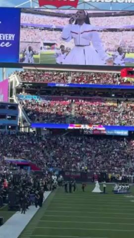 The video captures a live performance at a packed stadium, where a singer is visible on a large screen. The stadium is filled with spectators, who seem to be enjoying the performance. On the field, a small group of people are gathered near what appears to be a wedding party, including the bride in a white dress. People in suits are present. The overall atmosphere is lively and festive. The stadium has multiple tiers of seating and is illuminated by natural sunlight. The image on the screen shows the singer in a white outfit, accompanied by performers. The field is green.