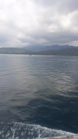 The video shows a wide expanse of calm, blue water with gentle ripples. In the distance, a small boat is visible on the water. Behind the boat, a lush, green coastline stretches out, backed by rolling hills and mountains under a cloudy sky. The perspective suggests the viewer is on another boat, looking towards the shore.