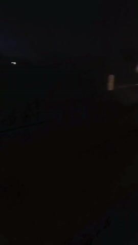 The video shows a dark, nighttime scene, possibly viewed from inside a vehicle. A bright light is visible in the distance, which appears to be a streetlight or a building light. The ground is dark and indistinct, with hints of what might be a road or path. There are no visible people or distinct objects other than the light source and a blurry reddish-orange shape in the far distance. The overall impression is one of low visibility and a dimly lit environment.