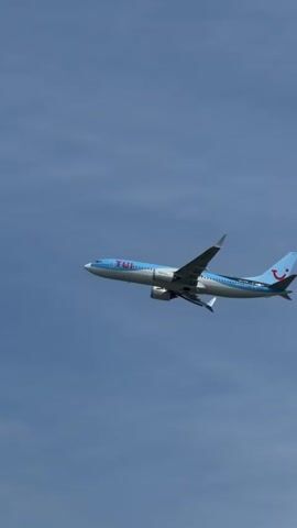 A TUI airline airplane is flying in a clear blue sky. The airplane is light blue and white with "TUI" written in red lettering on the side. The tail of the airplane is also light blue with a red logo. The airplane is shown from the side as it flies from left to right. Trees are visible at the bottom of the frame in the first few seconds. The camera is focused on the airplane.