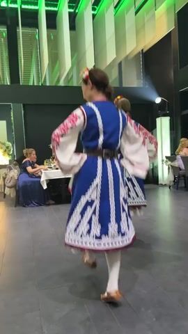 The video shows a group of people performing a traditional folk dance indoors. The dancers are dressed in colorful, elaborate costumes, with the women wearing blue and white dresses with intricate embroidery, and the men in white shirts, vests, and pants with dark accents. The dance appears to be energetic, with the performers moving in unison and at a brisk pace. In the background, guests are seated at tables, some of them observing the performance. The environment appears to be a banquet hall or reception area, with draped white fabric panels and green lighting visible on the ceiling. The floor is tiled. One dancer in the foreground is shown from behind, wearing a blue and white dress and white stockings. Another dancer in a similar costume moves past her to the left. A male dancer in a white costume with a black belt and hat enters the frame from the left, performing a dynamic move with his arm raised. The overall atmosphere suggests a festive occasion, possibly a wedding or cultural celebration.