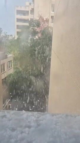 The video shows a view from a high window looking out at a hailstorm. Hail is accumulating on the window sill. Buildings and trees are visible in the background, partially obscured by the falling hail. The hail is dense and appears to be falling rapidly. The overall scene is somewhat obscured due to the intensity of the hail.