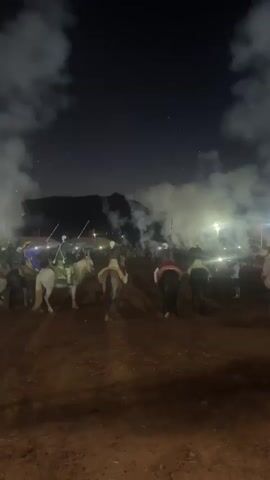 This media captures a nighttime event in Tabia, Morocco. The scene is filled with a dramatic atmosphere, dominated by thick smoke billowing across the dark sky. In the foreground, several individuals are mounted on horses, facing away from the camera and towards the center of the activity. They are dressed in what appears to be traditional attire, with some wearing military-style uniforms and hats.

The background is illuminated by bright lights, suggesting a gathering or festival. A prominent fire is visible in the distance, contributing to the smoke and adding a fiery orange glow to the scene. The ground is dirt, typical of an outdoor venue. The overall impression is one of a cultural or historical reenactment, possibly a fantasia, given the horses, riders, and pyrotechnics. The darkness of the night, combined with the smoke and fire, creates a sense of spectacle and perhaps excitement or intensity. There is no visible text in the media.