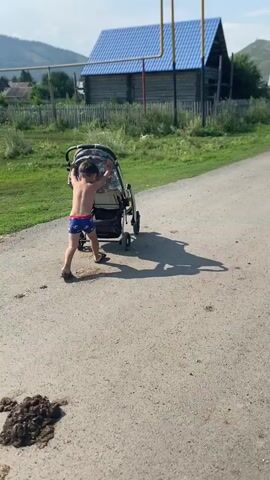 A young child, wearing red and blue striped shorts, pushes a baby stroller along a dirt road. The child is facing away from the camera, with their back to the viewer, and their arms are extended as they grip the stroller's handles. The child appears to be walking forward, pushing the stroller in front of them. The stroller is dark in color with a patterned fabric on the canopy and seat. The road is made of asphalt and is lined with grass on either side. In the background, there are wooden houses with blue roofs, a wooden fence, and rolling hills covered in trees. The sky is blue and sunny, with some clouds visible. The child's shadow is cast on the road behind them.