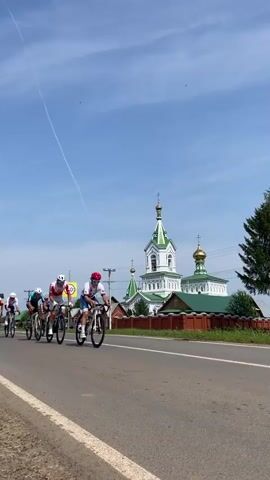 A group of cyclists race down a road with a church in the background. The cyclists are wearing racing gear and helmets. The church is white with a green roof and gold domes. The sky is blue with some clouds. The road is paved and there is a white line marking the edge of the road. There is a speed limit sign that says 20.