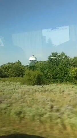 A horizontal video shows a view from a vehicle moving forward. In the distance, a white water tower is visible, surrounded by green trees. In the foreground, a grassy field is seen. The sky is clear and blue. Reflections from the vehicle's window are visible on the left side of the screen. The camera is moving from left to right, indicating the vehicle is traveling in that direction.