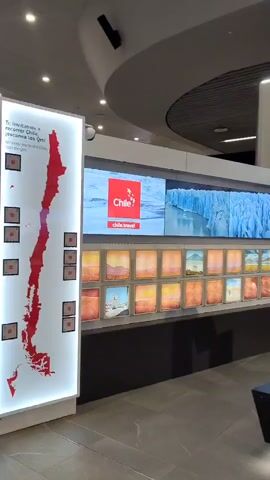 The video shows the Chile tourism booth in an airport or similar travel center. The word "Chile" is spelled out in large red letters, resting on a reflective black surface.  The word is punctuated by the Chile tourism logo and red stars hanging overhead. Behind the letters is a long wall covered in large screens playing videos of Chilean landscapes.  Below the video screens is a row of smaller framed photographs showcasing various scenes. To the left is a large red map of Chile highlighted in white.