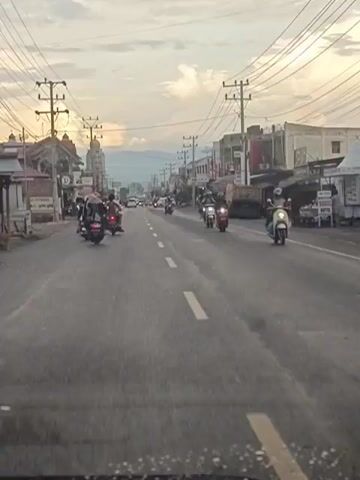 The media file captures a street scene in Barat Yaman, Indonesia, likely during the late afternoon or early evening. The sky is overcast with a soft, diffused light, suggesting either dawn or dusk. The road is asphalt, with a clear white dashed line marking the center, and a single yellow line running along the right side.

The primary subjects are numerous motorcycles and scooters navigating the road. Several individuals are riding these vehicles, some with passengers. The surrounding setting is a built-up area, with buildings lining both sides of the street. These structures appear to be shops or small businesses, indicated by signs and awnings. Power lines crisscross above the street, supported by utility poles, a common sight in many Indonesian towns. In the distance, mountains are faintly visible, silhouetted against the sky.

The activity is everyday traffic. Motorcycles are the dominant mode of transportation, and the scene suggests a moderately busy street. There are no obvious signs of a specific event; it appears to be a normal day.

Notable details include the presence of multiple utility poles and the general architecture of the buildings, which are indicative of the region. The lighting suggests a time of day when the sun is low on the horizon. No specific emotions are conveyed, as the footage is observational.

Visible text includes a circular sign with a 'D' on the left side of the street, and other illegible signs on the shopfronts. The overall impression is a typical street in a populated area of Indonesia, characterized by motor vehicle traffic and commercial establishments.