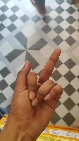 A close-up shot focuses on a person's hand against a patterned tile floor. The hand is positioned in the foreground, with the palm facing downwards and slightly towards the viewer. The index finger is extended upwards, while the middle finger is bent at the knuckle and touching the tip of the index finger. The thumb, ring finger, and pinky finger are curled inwards towards the palm. The skin tone of the hand appears to be medium brown, with short, natural fingernails. The background shows a geometric tiled floor with a checkerboard pattern of black and white squares, arranged in a radial design. In the upper portion of the frame, out of focus, are the lower legs and feet of at least one other person, wearing dark trousers and light-colored shoes. The lighting suggests an indoor environment, possibly a public space or a home.