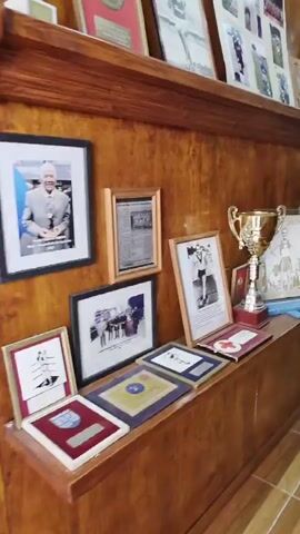 The video shows a wall display in what appears to be a club or museum, featuring framed photographs, newspaper clippings, and trophies.  The wall itself has wood paneling. Two shelves are lined with framed pictures and trophies of different sizes and colors.  A large glass display case houses a collection of black and white photos, likely documenting the history of the club or organization. The photographs showcase individuals and teams in various settings. The lighting in the room is adequate to illuminate the display.