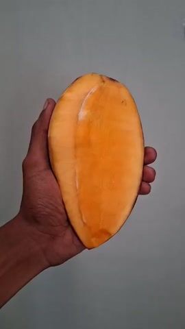 A hand holds a mango that is cut in half lengthwise. The inside of the mango is a bright orange color. The hand then turns the mango to show the other side, revealing a smaller piece of the mango that is still attached to the skin. The mango is then fully enclosed by the hand and rotated to show its entirety. The video then shows the hand holding the whole mango, which is a mix of yellow and green. The mango is then rotated to show its entirety. The hand is then shown holding the mango from a different angle. The mango is then rotated to show its entirety again.