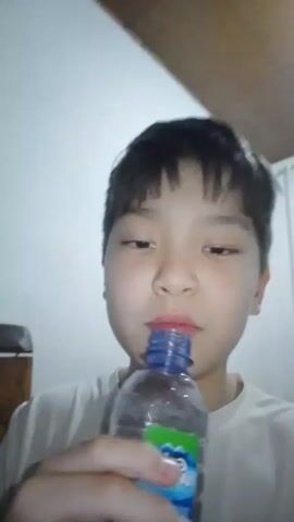 A young boy is drinking from a plastic water bottle. He is indoors in a room with white walls and a light wood ceiling. He has short, dark hair and is wearing a white t-shirt. He is looking at the camera while drinking.