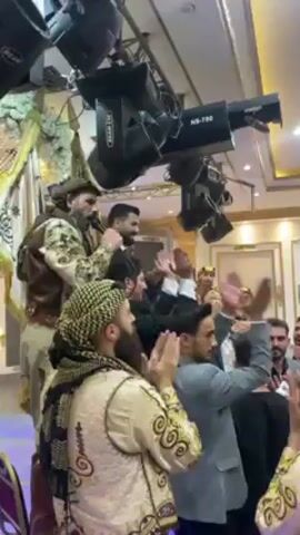 A group of people are gathered in what appears to be an indoor event space. The focal point of the scene is a man in a dark suit and tie, who is being lifted up amidst the crowd. Some individuals are clapping their hands, seemingly celebrating or showing support. Another man, wearing a camouflage jacket and headscarf, is holding a microphone and speaking. Several people are wearing traditional Middle Eastern clothing. Overhead, there are lighting fixtures and a ceiling decorated with gold accents. The room has a celebratory and festive atmosphere.