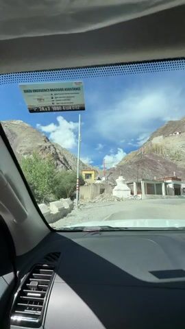 The video shows a view from inside a vehicle, looking through the windshield. The road is paved and appears to be in a mountainous region. On the left side of the road, there are several structures, some of which appear to be older buildings with stone walls and wooden doors and windows. A white stupa is also visible. Prayer flags are strung across some of the buildings and poles. The landscape is characterized by steep, rocky hillsides with sparse vegetation. The sky is blue with scattered white clouds. A sign for "ISUZU EMERGENCY ROADSIDE ASSISTANCE" is attached to a pole on the left side of the road. The vehicle is moving forward, suggesting the footage is from a car traveling along the road.