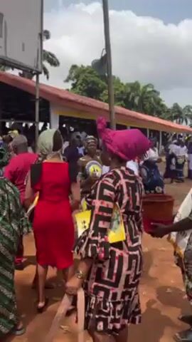 The video depicts a gathering of people outdoors on a partly cloudy day. Many individuals are dressed in colorful, traditional clothing. Some are dancing to music, while a man in white and purple clerical attire holds a collection bucket. There are structures with red roofs in the background, possibly tents or open-air buildings, suggesting a fair or celebration. A tall utility pole and various signs are also visible. The ground appears to be red dirt.