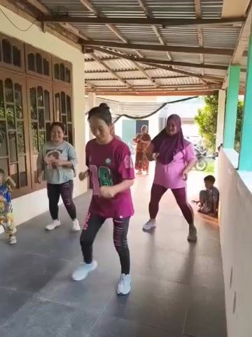 Four women and two children are seen in an outdoor covered walkway of a house. The women are dressed in athletic wear and appear to be exercising to music. The woman in the foreground wears a magenta shirt with the number 98 in green and black stripes. She is performing a dance move with her arms extended. To her left, another woman in a gray shirt and black leggings is also dancing. To her right, a woman in a pink hijab and matching pink leggings is also dancing. Further behind, another woman in a beige shirt can be seen. Two young children are present; one is on the far left wearing a colorful shirt and shorts, and another is sitting on the right, also in colorful clothing. The walkway has a gray tiled floor and a corrugated metal roof supported by wooden beams. Behind the walkway, part of the house is visible, including a large wooden-framed window. In the background, trees and a motorcycle are visible. The lighting suggests it is daytime.