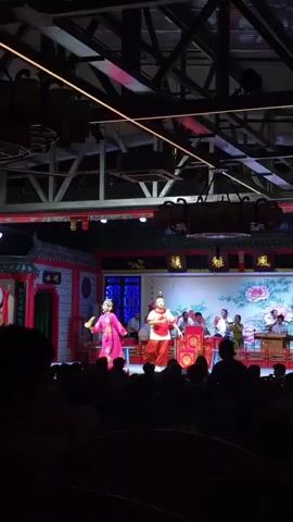 This is a wide shot of a stage performance. Two performers are on the stage. One is dressed in a bright pink dress and the other in a white and red outfit with a headpiece. There are musicians playing instruments behind them. The stage is decorated with traditional Chinese architecture and a backdrop of peonies. The audience is visible in the foreground, silhouetted against the stage lights. The overall atmosphere appears to be a traditional Chinese opera or theatrical performance.