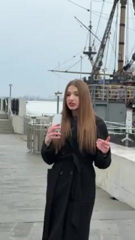 A young woman with long brown hair is standing on a paved walkway, speaking into her smartphone. She is wearing a long black belted coat. Behind her is a large, old-fashioned sailing ship with tall masts and rigging. To the left of the woman are gray concrete steps and a metal railing. The sky is overcast, and the background suggests a waterfront location. The woman gestures with her hands as she speaks, indicating the ship behind her.