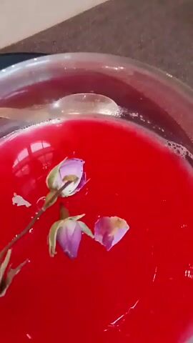 A close-up shot shows a small glass bowl filled with a bright red liquid, decorated with a small rose stem and petals. The bowl sits on a black plate. The camera zooms out to reveal the setting: a restaurant with tables, chairs, and a white ramekin nearby. The restaurant has a formal ambiance.