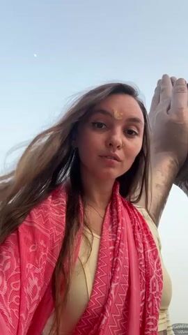 A woman stands outdoors in front of a statue of two hands in a praying pose. She is wearing a yellow shirt and a pink scarf with a white pattern. Her brown hair is blowing in the wind, and she has a gold mark on her forehead. The sky is pale blue, and there is a small crescent moon visible. The background includes a bridge and some people, slightly blurred. The woman looks directly at the camera with a slight smile.