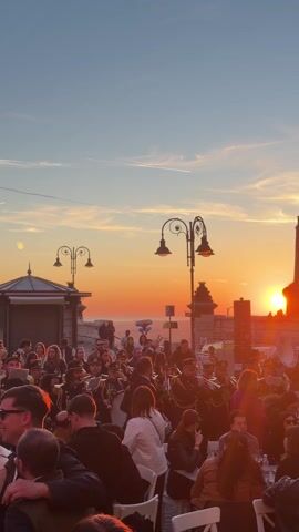The video captures a vibrant scene at sunset. A large group of people is gathered in an outdoor area, possibly a square or terrace. A marching band in uniform is performing, with musicians playing brass instruments. The crowd is a mix of individuals, some standing and watching the band, while others are seated at tables, enjoying drinks and conversation. The sun is setting in the background, creating a warm, orange glow in the sky. There are street lamps and building structures in the background as well. The scene conveys a lively, festive atmosphere.