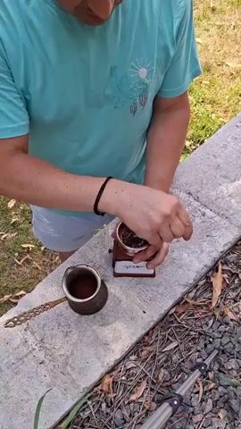A person wearing a turquoise t-shirt and white shorts is standing outdoors, leaning over a concrete ledge. They are using a small, manual coffee grinder with a copper-colored exterior. The person is holding a small metal tool, possibly a spoon or stir stick, and is manipulating the contents of the grinder. Beside the grinder is a small copper coffee pot, known as a cezve or ibrik, containing a dark liquid. The background shows dry leaves on the ground and some green grass.